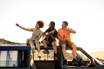 Fototapeta na wymiar A group of 3 young multi-ethnic people are having fun on top of a camper van at sunset. Friends are taking a picture with the mobile phone while drinking beer and playing a flamenco guitar.