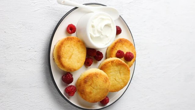 Baked keto syrniki on a ceramic plate with sour cream and raspberries, a female hand twists the plate on a light gray background