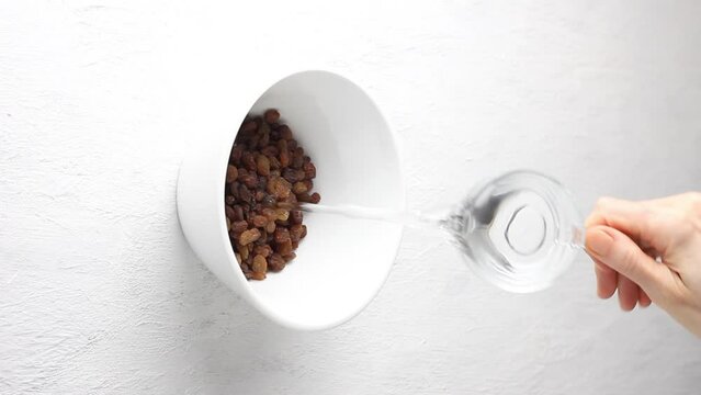 Raisins are poured into a white bowl and filled with water for soaking and rinsing on a gray textured background