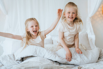 Excited sisters laughing sitting on bed celebrating great news. Screaming blonde preschooler girls...