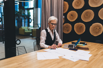 Serious aged businesswoman working with documents and laptop in office