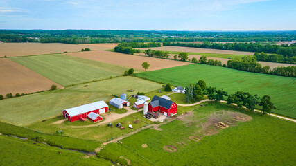 Green pastures and farmland around classic red barn with tractors and distant dirt fields aerial