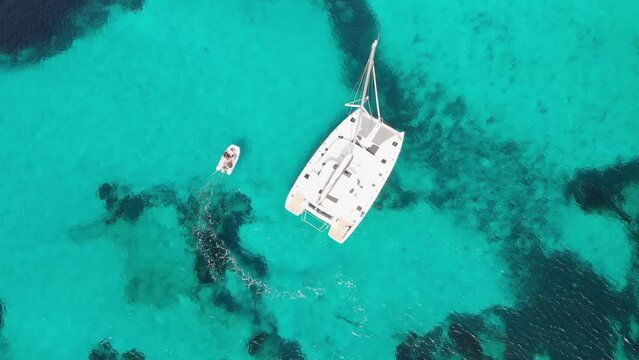 aerial stunning drone view of a dinghy leaving its trail floating around a catamaran in a crystal clear turquoise sea