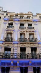 Fototapeta na wymiar Belle façade d'un batiement parisien de luxe ou hotel chic, sous un beau jour, avec de jolis projecteurs bleus, beauté urbaine, immeuble en ancien pierre, style riche, à plusieurs étages, balcon