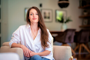 Obraz premium Attractive brunette haired woman wearing white shirt and blue jeans and relaxing in an armchair at home
