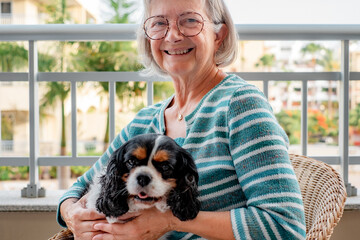 Portrait of senior woman sitting on the home balcony holding her cavalier king charles dog on the...