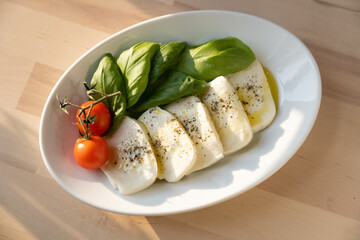 Fresh mozzarella, tomatoes, and sweet Italian basil on a white plate next to a kitchen window at sunset 