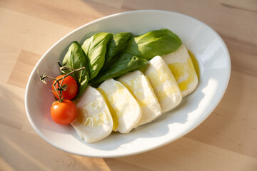 Fresh mozzarella, tomatoes, and sweet Italian basil on a white plate next to a kitchen window at sunset 