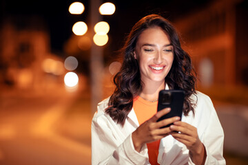 Lifestyle portrait of happy woman walking in evening city and using cellphone, texting or surfing internet, free space