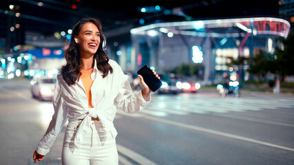 Fototapeta premium Trendy happy woman listening favorite music in earphones and dancing, walking in the evening on city street, free space