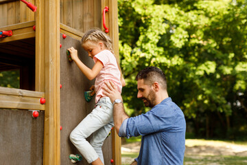 Father and daughter walking in park, girl trying to climb on wall at playground, family spending time together outdoors