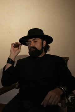 A cheeky strict bearded man in a black suit and black hat sits in a vintage armchair and smokes a cigar. Vertical photo.