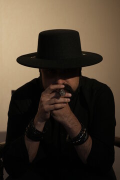 An adult bearded dark-skinned man in a black shirt and hat smokes a cigar thoughtfully. A serious business man sits on a chair and takes a puff on his cigar. Portrait. Vertical photo.