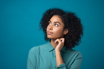 Fototapeta na wymiar Headshot portrait of thoughtful pensive young black hair woman with curly hair holding finger on lips looking upward against turquoise studio wall background with copy space for text advertisement