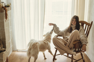 Happy woman in cozy knitted sweater playing with dog while sitting in vintage wooden chair. Cozy...