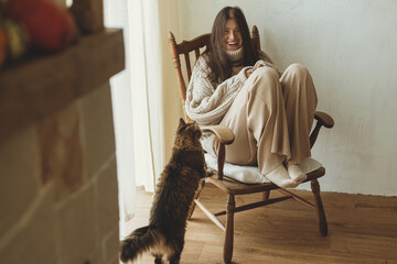 Happy woman in cozy knitted sweater playing with cat while sitting in vintage wooden chair. Cozy...