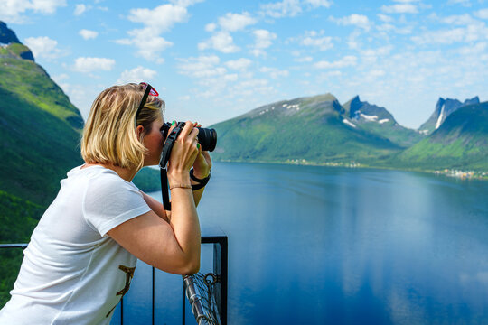Young woman photographer making landscape photograpy of Norway fjords in bright summer day. Young woman with standing on a hill with green grass and making snapshot of a beautiful scene, arial view