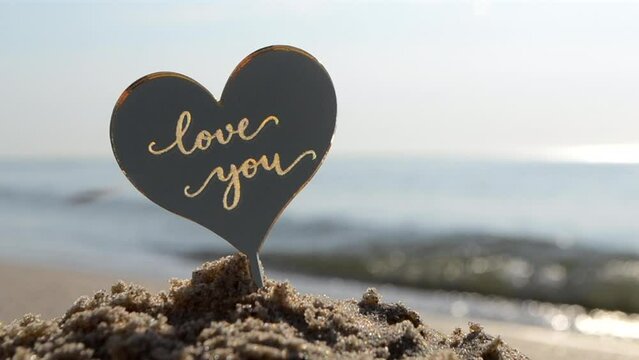Plastic stick shape of heart with words Love you in sand beach of sea shore on background sea waves on sunny summer day close-up. Concept love relationship romance amour celebration St. Valentines Day