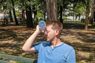 Man suffering from heat, guy with heatstroke. Caucasian male presses bottle of water to face to cool off suffering from heat wave, stuffiness - obrazy, fototapety, plakaty