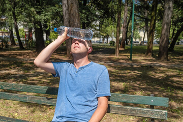 Man suffering from heat wave Charon in South Europe. Caucasian male presses bottle of water to face to cool off suffering from heat sitting outdoors - obrazy, fototapety, plakaty