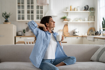 Happy dreamy young woman wearing casual clothes resting with cup of tea at cozy home, smiling female sitting on couch enjoying favorite hot drink, sipping it slowly to relax. Hygge and slow life - obrazy, fototapety, plakaty