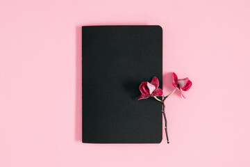 Flat lay, top view of blank black notebook mockup and orchid flowers on pastel pink background