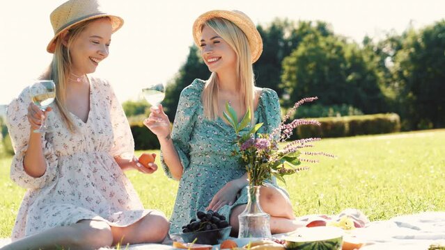 Two young beautiful smiling hipster female in summer sundress and hats. Carefree women making picnic outside. Positive models sitting on plaid on grass, drinking wine, eating fruits. Cheerful, happy