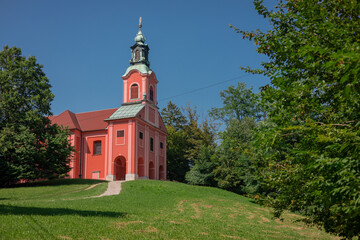 Red church on Rozik hill above Ljubljana surrounded by lush green trees and grassy meadows. Beautiful scenery around a tyoical church.