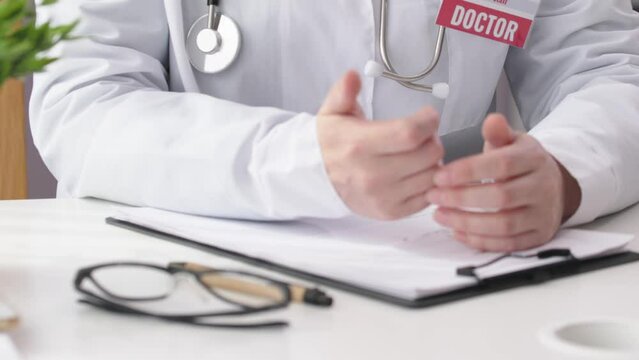 unrecognizable doctor hands in white coat sit table gesturing talking in office. concept doctor's appointment prescriptions medicine worker speech. white robe, stethoscope, glasses, clipboard on desk