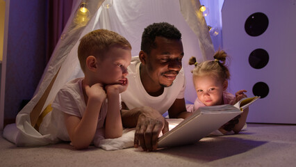 African american man babysitter reads book to caucasian kids in playroom home.