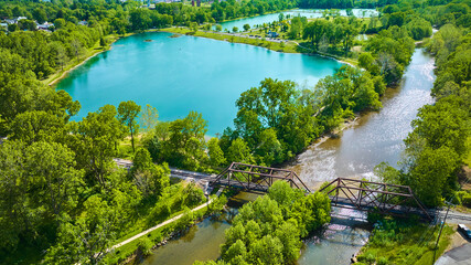 Fototapeta na wymiar Aerial beautiful teal blue pond with summertime trees and a river running under a train bridge