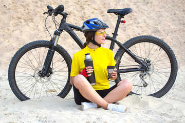 Beautiful woman cyclist in yellow with tea and thermos in hand on the background of sand. Sports and recreation.