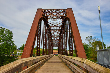 Brown iron walking bridge with metal crisscrossing beams under a cloudy blue sky
