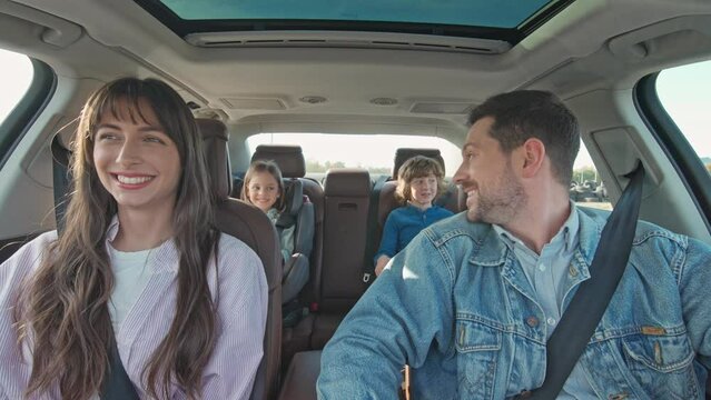 Cheerful young traditional family has long auto journey together. Parents and two children going on family trip. Safety riding car concept wide angle inside car view image.