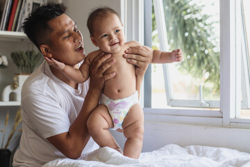 Happy excited young father and his baby playing together near window in the bedroom. Fatherhood,...