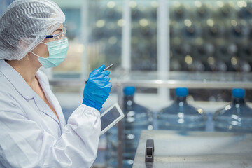 scientist worker checking the quality of water bottles on the machine conveyor line at the industrial factory. Female worker recording data at the beverages manufacturing line production.