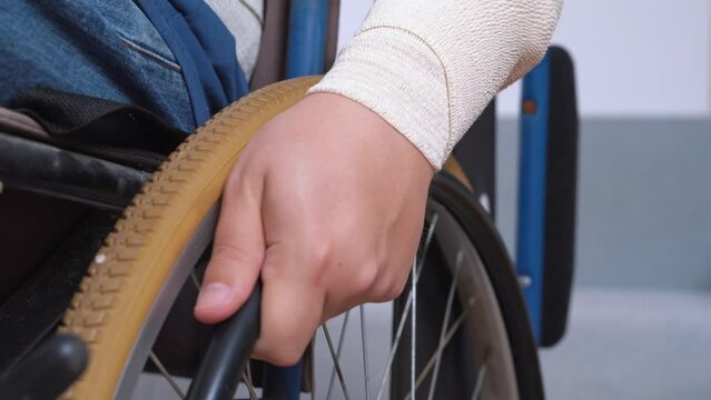 Wheel of mechanical wheelchair, man in jeans with bandaged hand twists, moves forward back indoors Close-up. Patient injured after accident, in cast, moves mobility aid device. Independent, disability
