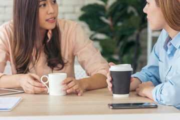 Two Women colleagues drinking black coffee cup at office desk together. Asian woman friendstalking smiling with friendship happy time. Teamwork happy moment lifestyle. International Woman day concept