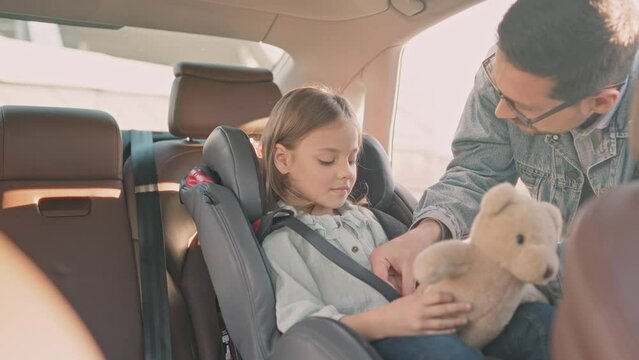 Adult man securing little daughter in car seat in his vehicle. Cheerful young handsome father on trip in car with teen daughter touching her nose to cheer up. Childhood and parenting, travel concept.