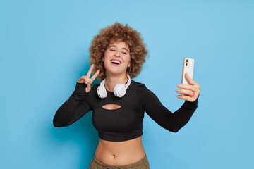 Pretty girl with short afro hair in black longsleeve top taking selfie on blue background, laughing...