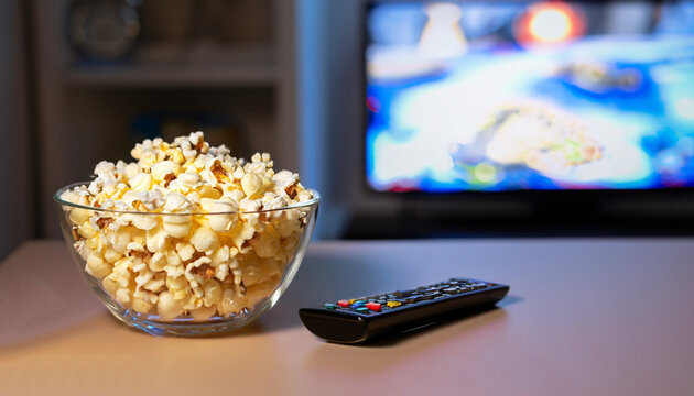A glass bowl of popcorn and remote control in the background the TV works. Evening cozy watching a movie or TV series at home - Powered by Adobe