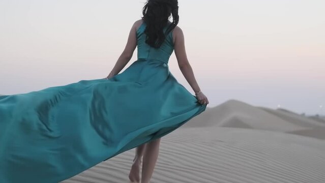 Young woman with black hair walking barefoot in a long blue evening dress on the sand dunes of the desert