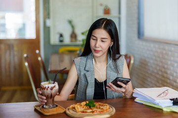 Obraz na płótnie Canvas Asian beautiful woman sitting in a small restaurant Preparing to eat lunch alone Order pizza and coffee to eat. along with listening to music via smartphones for enjoyment