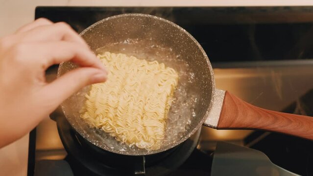 POV Hands Cooking Instant Noodle Soup At Home. Female Hand Putting Dry Noodles Into Boiling Water In A Pan At Home And Stirring. Delicious And Simple Snack. Student Life. Steaming Food On Gas Stove.