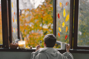 A cute boy wrapped in a blanket drink hot tea and looks out the open window at the wonderful autumn...
