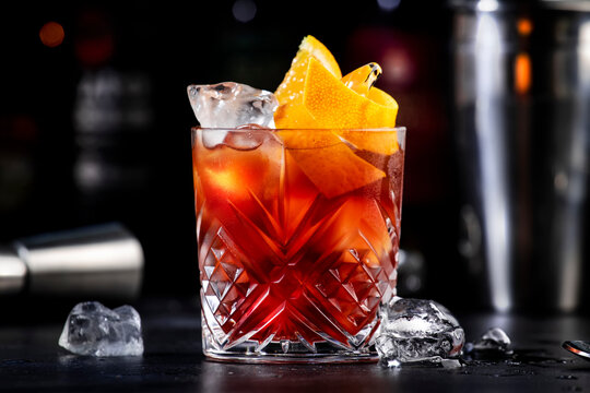 Espresso Negroni alcoholic cocktail drink with dry gin, red vermouth and red bitter, espresso and coffee liqueur, orange slice and ice cubes. Black bar counter background, bar tools, bottles