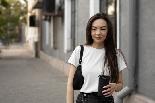 Attractive Asian long-haired brunette woman dressed in white t-shirt with black leather bag and coffee to-go walking along the street