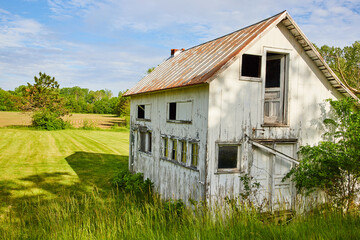 Fototapeta na wymiar Abandoned farm house in grassy field near farmland with busted windows and door