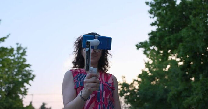 Young woman with glasses testing innovative new Insta360 flow mobile camera gimbal stabilizer at sunset. Low angle