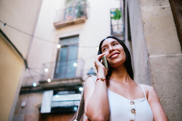 From below glad Latin American female smiling and looking away while leaning on wall of apartment building and answering phone call on street of Barcelona, Spain